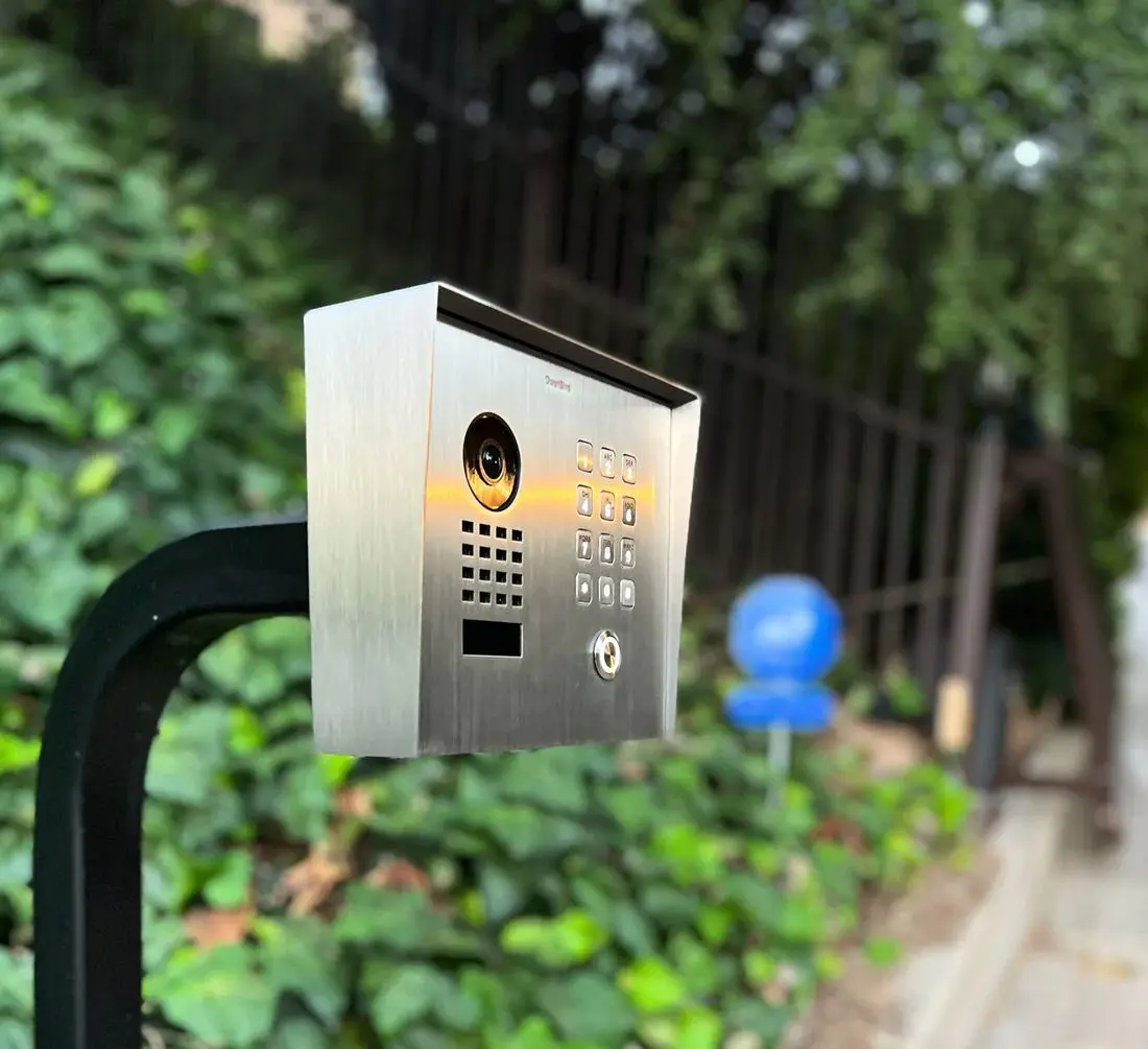 DoorBird video intercom with keypad mounted on a gooseneck post at a gate